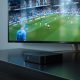 Android TV streaming box resting on a matte dark wooden surface below a flat-screen television displaying a football match broadcast, red warning icon subtly visible on screen edge, shot at 45-degree angle using a 50mm lens, soft natural window light from the left, realistic surface reflections, no hands, no people, no text watermarks, photorealistic, 16:9