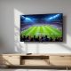 A Samsung smart TV mounted on a light grey living room wall, screen displaying a live football match broadcast with a stadium crowd visible, a small streaming remote resting on a wooden console below, shot at a 45-degree angle using a 50mm lens, soft natural window light from the left casting long diffused shadows, realistic screen reflections, shallow depth of field, no hands, no people, no text overlays, no watermark, photorealistic, 16:9