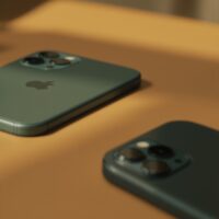 iPhone 11 Pro in matte midnight green resting slightly out of focus on a warm beige surface, with a newer iPhone 17 Pro in sharp focus in the foreground on the same surface, soft cinematic side lighting with subtle directional shadows, no people, no hands, no text, photorealistic, 16:9