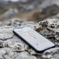 iPhone 18 Pro resting on a rocky outdoor stone surface with rugged terrain blurred in the background, screen showing a Maps navigation interface with a route active, shot at 45-degree angle using a 50mm lens, soft overcast natural light from the left, shallow depth of field, no hands, no people, no text, no watermark, sharp focus, photorealistic, 16:9