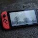 Nintendo Switch 2 console in handheld mode resting on a rough dark concrete surface, screen showing a misty post-Soviet landscape with grey skies and sparse trees, 45-degree angle, 50mm lens, shallow depth of field, soft natural window light from the left, long diffused shadows, realistic reflections on glossy console housing, no hands, no people, no text, no watermark, sharp focus, photorealistic, 16:9